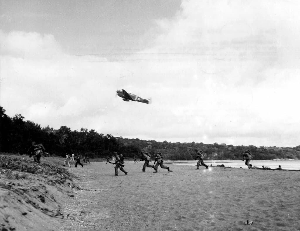 Landing operations on Rendova Island, Solomon Islands, beginning 30 June 1943.  Flying at 'tree-top height a U.S. Army P-40 snarls over a group of American troops chasing across the beach.